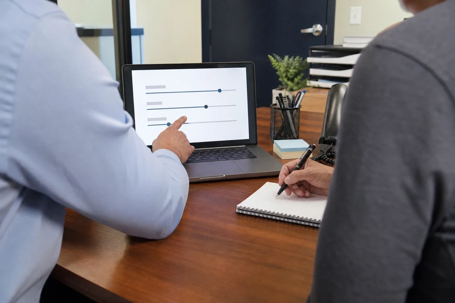 Over-the-shoulder view of an agent pointing at slider options on a laptop while a client takes notes in a spiral notebook at an office desk